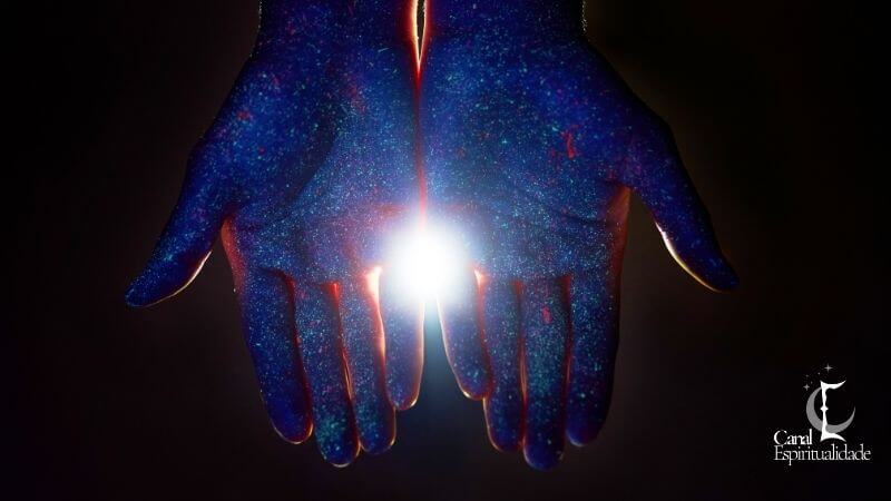Fotografia Kirlian - Efeito Corona Duas mãos abertas voltadas para cima em um ambiente escuro. Elas estão cobertas por uma textura brilhante que lembra poeira estelar ou galáxias em tons de azul, ciano e pontos avermelhados. No centro, entre as palmas, há uma forte fonte de luz branca radiante que ilumina o interior das mãos.