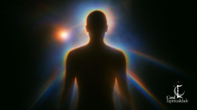 Fotografia Kirlian - estado emocional e o fluxo vital Silhueta escura do tronco e cabeça de uma pessoa contra um fundo preto. O contorno do corpo é suavemente iluminado por uma aura gradiente que vai do laranja ao azul. Ao redor da cabeça e dos ombros, há halos de luz colorida e reflexos em formato de arco-íris, sugerindo um campo vibracional.