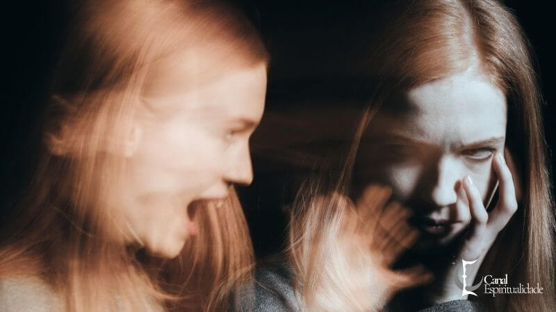 Fotografia em estilo de exposição múltipla ou sobreposição de imagem de uma mulher com longos cabelos ruivos, dividida em dois estados emocionais distintos em um fundo escuro. Do lado esquerdo, a imagem desfocada e em movimento mostra a mulher com a boca aberta em um grito, com uma expressão de raiva ou angústia. Do lado direito, a imagem mais nítida mostra a mulher com a mão no rosto, cobrindo a boca e olhando para baixo com uma expressão de desespero ou tristeza profunda, com a pele pálida e os olhos escuros. A imagem simboliza a luta interna e o sofrimento emocional. No canto inferior direito, há um logotipo roxo com o texto "Canal Espiritualidade".